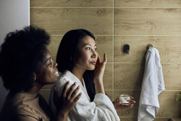 Smiling woman looking at girlfriend applying moisturizer on face in bathroom - DSHF00492