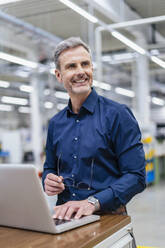 Lächelnder Geschäftsmann mit Laptop in einer Fabrik - DIGF18003