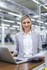 Portrait of smiling businesswoman with laptop and documents in factory - DIGF18013