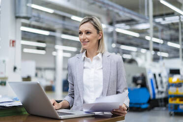 Smiling businesswoman with laptop and documents in factory - DIGF18015