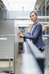 Geschäftsfrau lehnt an einem Geländer in einer Fabrik und benutzt ein digitales Tablet - DIGF18044