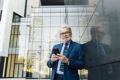 Smiling businessman standing with smart phone by wall outside office building - MEUF06245