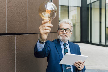 Businessman with light bulb and tablet PC in hand standing outside office building - MEUF06250