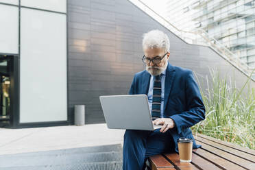 Senior businessman working on laptop on bench outside office park - MEUF06255