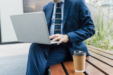 Geschäftsmann, der an einem Laptop arbeitet, während er auf einer Bank im Büropark sitzt und eine Tasse Kaffee trinkt - MEUF06256