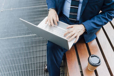 Businessman working on laptop sitting on bench by coffee outdoors - MEUF06257