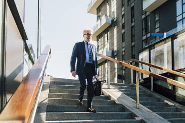 Älterer Geschäftsmann mit Aktentasche geht an einem sonnigen Tag die Treppe hinunter - MEUF06270