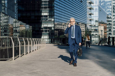 Älterer Geschäftsmann, der mit seinem Smartphone spricht und vor einem Bürogebäude geht - MEUF06275