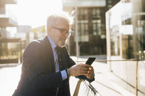 Älterer Geschäftsmann mit Smartphone am Geländer stehend an einem sonnigen Tag - MEUF06277