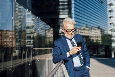 Älterer Geschäftsmann, der an einem Smartphone arbeitet und vor einem Bürogebäude steht - MEUF06278