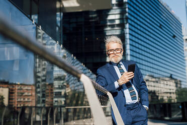 Smiling businessman standing with smart phone outside office building on sunny day - MEUF06279
