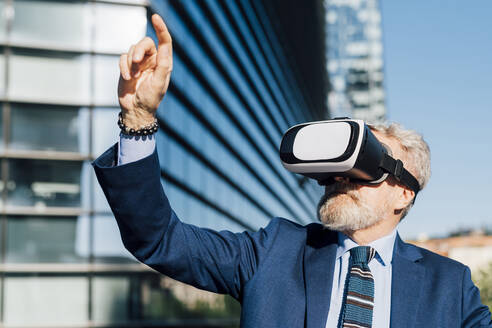 Älterer Geschäftsmann mit Virtual-Reality-Headset gestikuliert vor einem Bürogebäude - MEUF06284