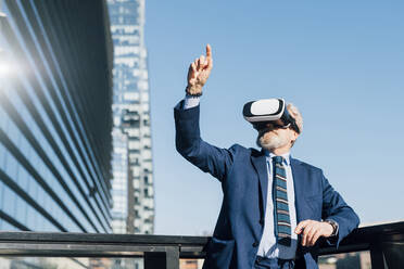 Ein älterer Geschäftsmann zeigt mit Gesten auf einen Virtual-Reality-Simulator außerhalb eines Bürogebäudes - MEUF06285