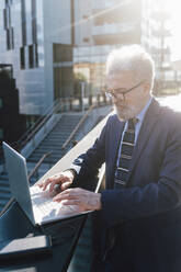 Ein älterer Geschäftsmann arbeitet an einem Laptop mit einem Solar-Ladegerät vor einem Bürogebäude - MEUF06303