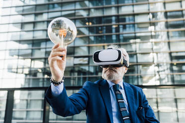 Senior businessman with virtual reality simulator holding light bulb outside office building - MEUF06307