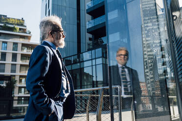 Älterer Geschäftsmann mit Hand in der Tasche, der das Glas eines modernen Gebäudes betrachtet - MEUF06311