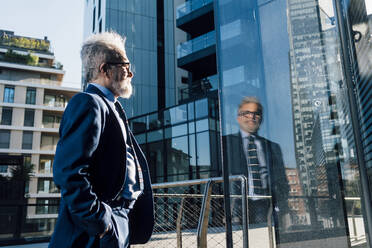 Senior businessman with hand in pocket looking at glass of modern building - MEUF06311