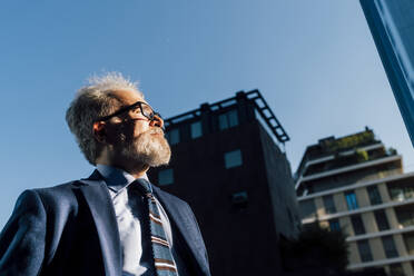 Senior businessman looking away standing outdoors on sunny day - MEUF06312