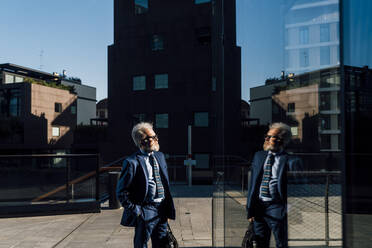 Ein älterer Geschäftsmann steht mit den Händen in den Taschen vor einem Bürogebäude an einem sonnigen Tag - MEUF06313