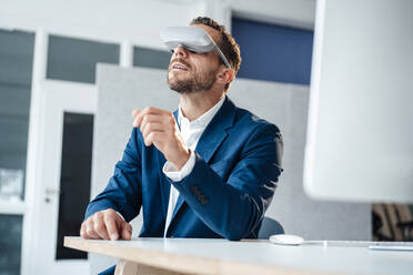 Businessman wearing virtual reality headset sitting at desk in office - MOEF04137