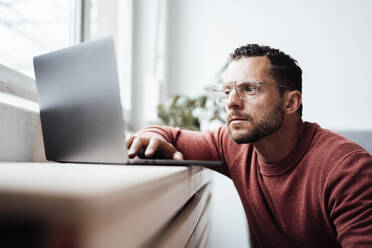 Freiberufler mit Brille arbeitet zu Hause am Laptop - MOEF04139