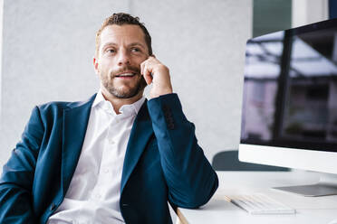 Lächelnder Geschäftsmann, der am Schreibtisch im Büro sitzt und mit seinem Smartphone spricht - MOEF04140