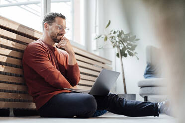 Smiling freelancer looking at laptop sitting on floor at home - MOEF04143