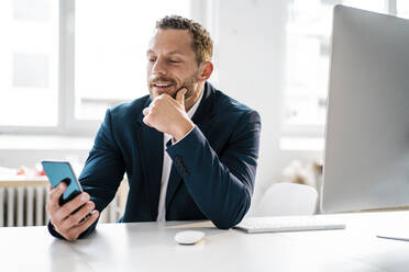 Smiling businessman with hand on chin using smart phone in office - MOEF04144