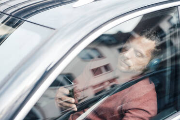 Mann mit Kopfhörern und Smartphone im Auto sitzend durch Fenster gesehen - MOEF04148