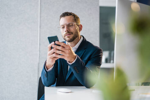 Lächelnder Geschäftsmann, der ein Smartphone benutzt und am Schreibtisch im Büro sitzt - MOEF04149