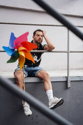 Man with head in hand holding pinwheel toy sitting near railing - MOEF04156