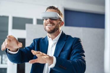 Happy businessman in virtual reality headset gesturing in office - MOEF04165