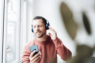 Man holding smart phone enjoying music on headphones at home - MOEF04169