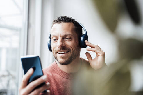 Lächelnder Mann mit Kopfhörern, der zu Hause sein Smartphone benutzt - MOEF04170