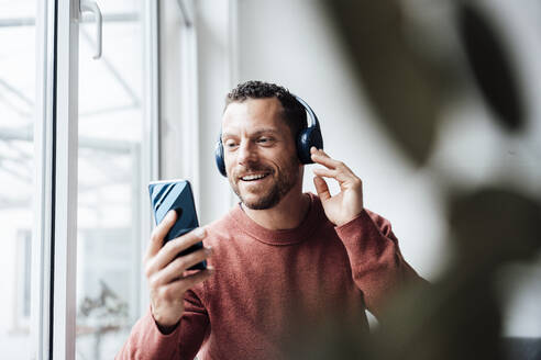 Lächelnder Mann hört Musik über Kopfhörer und hält sein Smartphone zu Hause - MOEF04173