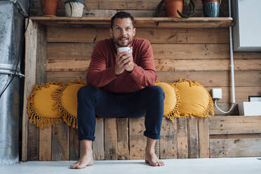 Smiling man holding cup sitting by cushions on bench - MOEF04177