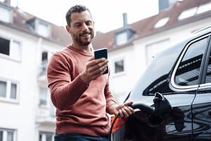 Lächelnder Mann, der ein Smartphone benutzt und ein Elektroauto vor einem Gebäude auflädt - MOEF04189