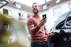 Glücklicher Mann mit Smartphone, der sein Elektroauto an einer Tankstelle auflädt - MOEF04193