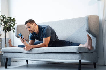 Mann, der sein Smartphone benutzt, sitzt breitbeinig auf dem Sofa zu Hause - MOEF04213