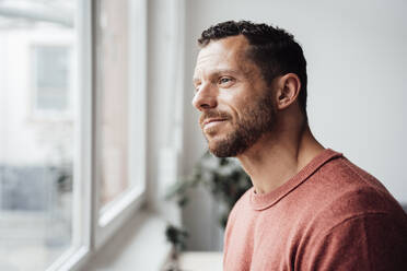 Lächelnder Mann schaut zu Hause aus dem Fenster - MOEF04240