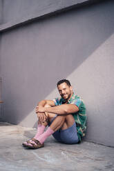 Smiling man sitting on floor near wall - MOEF04263