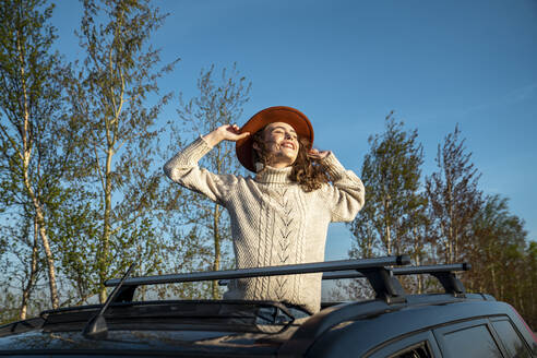 Glückliche Frau mit Hut steht mit geschlossenen Augen durch das Sonnendach eines Autos - VPIF06352