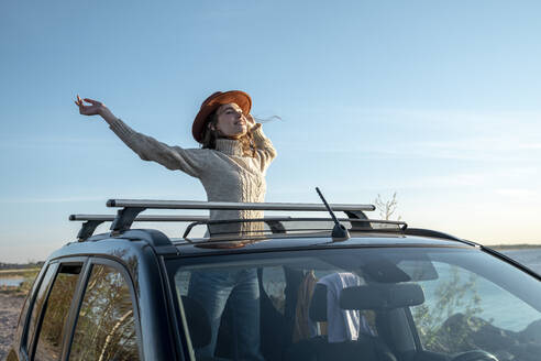 Glückliche Frau mit Hut steht mit erhobener Hand durch das Sonnendach eines Autos - VPIF06354