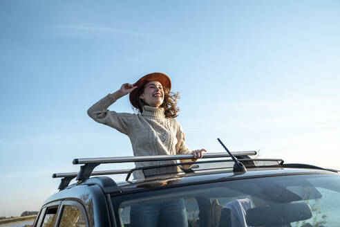Fröhliche junge Frau mit Hut, die durch das Sonnendach eines Autos genießt - VPIF06355