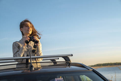 Lächelnde junge Frau mit Fernglas, die auf dem Sonnendach eines Autos steht - VPIF06359