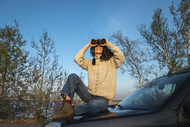 Glückliche Frau, die auf der Motorhaube sitzend durch ein Fernglas schaut - VPIF06385