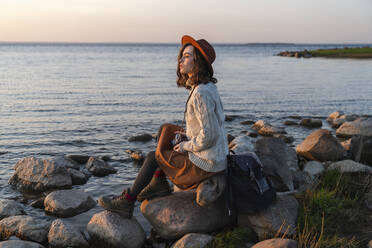 Young woman wearing hat sitting on rock at sea coast - VPIF06390