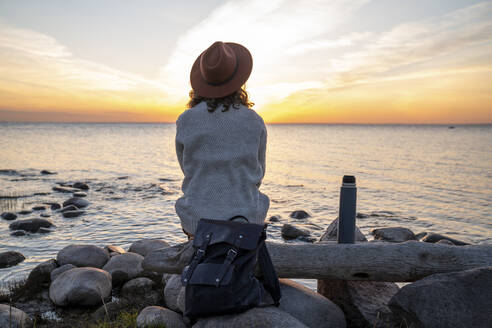 Junge Frau mit Rucksack auf einem Baumstamm sitzend bei Sonnenuntergang - VPIF06398