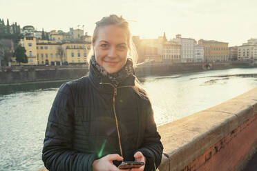 Smiling young woman with mobile phone standing by wall on sunny day - TAMF03428