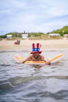 Lächelnder Mann mit Onkel-Sam-Hut schwimmt mit einem Pool-Floß im Meer - OCMF02446